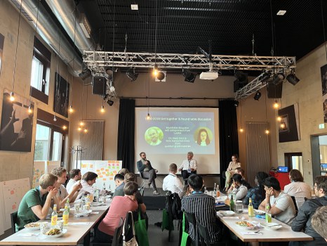 Get-together & Rundertisch-Diskussion mit 2 externen Speakern, Teilnehmende sitzen an zwei langen Tischen und hören gespannt zu