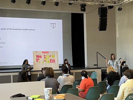 Workshop von einer der Trainerinnen, 2 Teilnehmende präsentieren ihr Board, die restlichen Teilnehmenden sitzen als Publikum