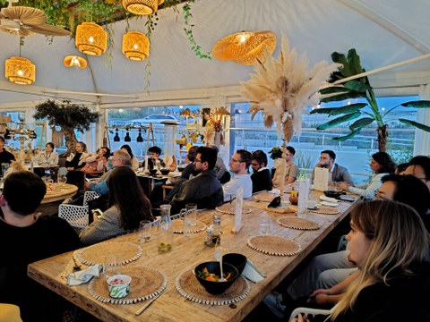 Viele Personen sitzen in einem Restaurant und hören drei Vortragenden zu