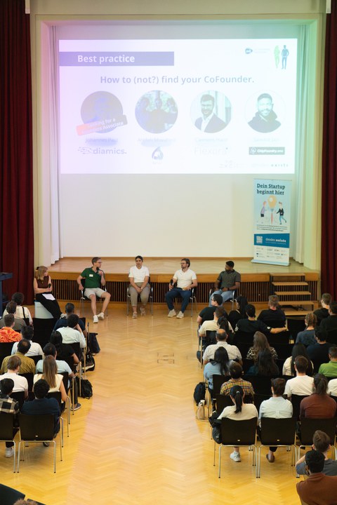 vor Bühne sitzen vier Personen, die im Panel diskutieren; davor sitzt Publikum 