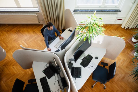 Drei Büroarbeitsplätze von oben fotografiert, ein Mann arbeitet gerade auf einem Platz am Computer.