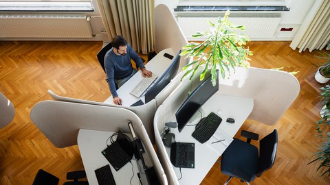 Drei Büroarbeitsplätze von oben fotografiert, ein Mann arbeitet gerade auf einem Platz am Computer.