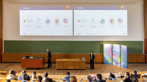 Foto eines Hörsaals mit mehreren Personen auf den Sitzplätzen. Vorn stehen zwei Personen und an der Tafel ist eine Präsentation zu sehen. 