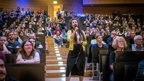 Blick in einen vollen Hörsaal. Im Gang steht eine Frau, die ins Mikrofon spricht.