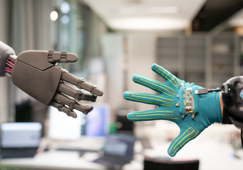 The photo shows two hands: a robotic hand and a human hand wearing a glove with electrical sensors.