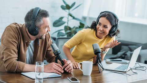 Foto von zwei Personen mit Kopfhörern, die in einem Studio sitzen. Vor ihnen stehen Mikrofone und ein Laptop.