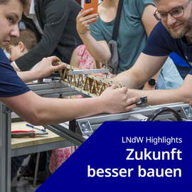 Foto: 2 Personen stehen sich gegenüber und halten zwischen sich ein Holz-Modell einer Brücke auf einem Tisch mit Werkzeugen im Hintergrund