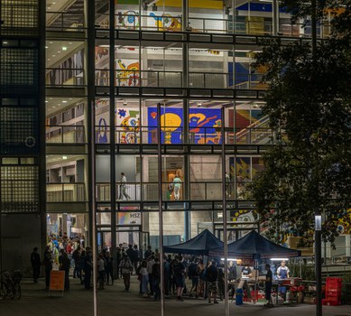 Foto des Hörsaalzentrums der TU Dresden am Abend mit Blick ins Innere. Es sind bunte Wände zu sehen und viele Personen vor dem Gebäude. 