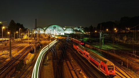 Das Foto zeigt viele Bahnschienen. Auf einer erkennt man einen roten Zug, auf einer anderen lediglich helle Lichtstreifen. Im Hintergrund befindet sich der hell erleuchtete Dresdner Hauptbahnhof.