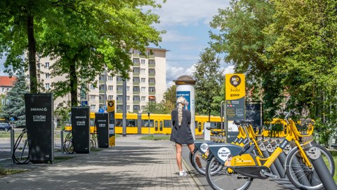 Foto einer Frau umgeben von "Drewag Stromtankstellen" links und "MOBI bike" Leihräder rechts. Im Hintergrund fährt die Straßenbahn vorbei.
