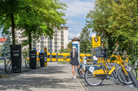 Foto einer Frau umgeben von "Drewag Stromtankstellen" links und "MOBI bike" Leihräder rechts. Im Hintergrund fährt die Straßenbahn vorbei.