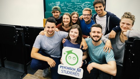 Gruppe junger Studierender im Hörsaal halten das Logo von "Psychisch fit Studieren" in den Händen