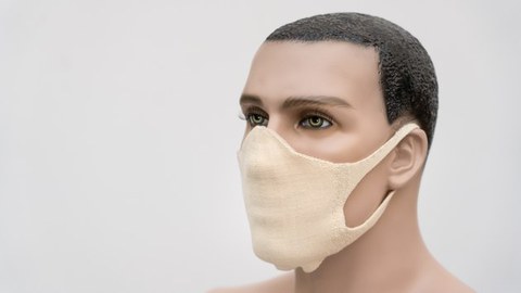 Gesicht einer männlichen Schaufesnterpuppe, deren Gesicht von Nase bis Kinn von einer Maske aus Stoff bedeckt ist, die an Bändern hinter den Ohren liegt.