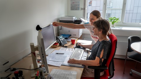 Eine Schülerin des physiotherapeutischen Lehrzentrums zeigt einer Mitarbeiterin  am Schreibtisch den richtigen Abstand zum Bildschirm