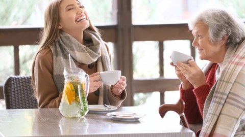 Foto einer jungen und älteren Person an einem Tisch beim Kaffee trinken. Beide sind fröhlich.
