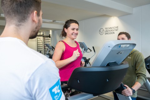 Frau mit pinkem Top auf Laufband wird von 2 Trainern beraten