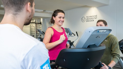 Frau mit pinkem Top auf Laufband wird von 2 Trainern beraten