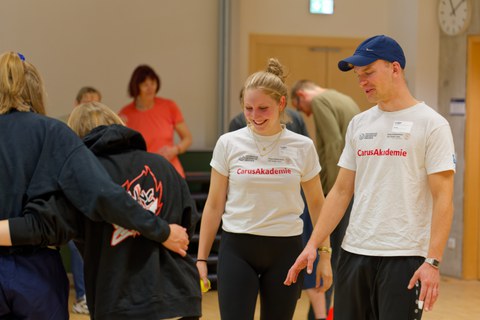 Zwei fröhliche Schüler:innen, welche die Bewegungsausführung von Teilnehmenden eines Bewegungskurses kontrollieren