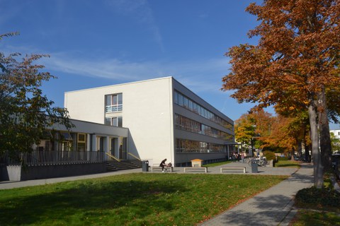 Eine Studentin sitzt auf der Bank vor dem Gebäude auf der August-Bebel-Straße. Ein Baum mit herbstlichen Blättern steht vor dem Gebäude.