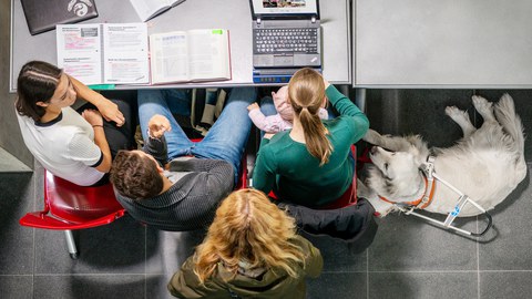 Vier Personen aus der Vogelperspektive aufgenommen. Drei davon sitzen nebeneinander an einem Schreibtisch. Die vierte Person steht hinter ihnen und blickt ebenfalls auf den Schreibtisch. Die rechtssitzende Studentin hält ein Kleinkind auf ihrem Schoß, ihre rechte Hand liegt auf der Braillezeile eines Notebooks, das aufgeklappt auf dem Tisch steht. Rechts von ihr liegt ein Blindenführhund. Auf dem Tisch liegen Bücher, ein Spiralblock, ein zugeklapptes Notebook und Kopfhörer.