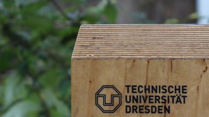 Zu sehen ist das Logo der TU Dresden auf einem Holzbrett. Im Hintergrund erkennt man verschwommen das Grün von Bäumen.