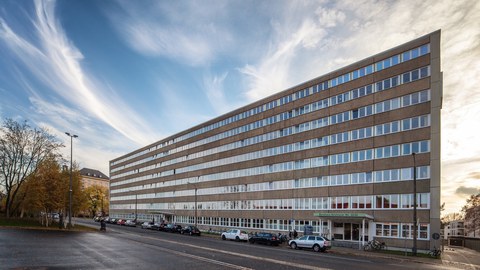 Das Bürogebäude Strehlener Straße mit seiner langen Fensterfront. Es liegt direkt an der Straße. Zu sehen sind viele Fahrräder, Autos, ein Kinderwagen, Bäume und ein wolkenverhangener Himmel.