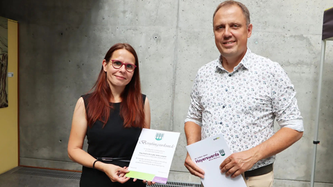 Eine Frau und ein Mann stehen nebeneinander vor einer Betonwand. Prof. Anke Langner mit langen kastanienbraunen Haaren und Brille hält eine Berufungsurkunde. Bürgermeister Mirko Pink im hellen geblümten Hemd hält eine Mappe mit der Aufschrift "Wir lieben Ideen. Hoyerswerda"