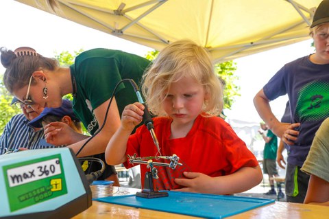 Ein Kleinkind mit blonden langen Haaren arbeitet mit Lötkolben an einem Stand bei den Zukunfstagen Hirschfelde