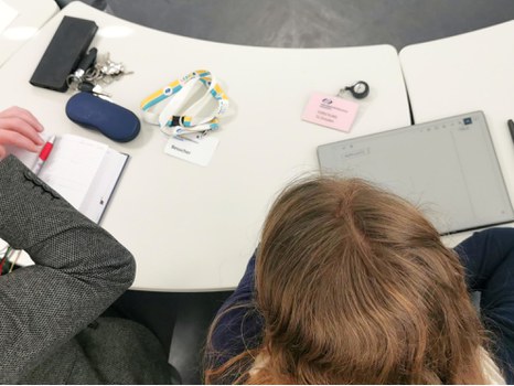 Blick von oben auf einen Konferenztisch mit Tablet und Schreibsachen, daneben ein Besucherschild "Universitätsschule Dresden" und "Forschungsstelle Universitätsschule ForUS"