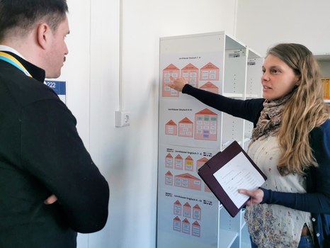 Eine Projektmitarbeiterin erklärt einem Schulleiter die sogenannten Lernhäuser auf einem Plakat in der Materialbibliothek der Universitätsschule Dresden.
