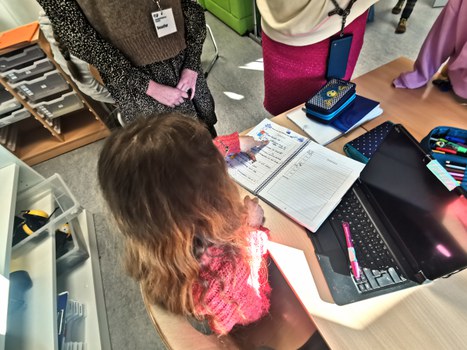 Blick von oben auf einen Schreibtisch. Eine Schülerin zeigt auf die Eintragungen in ihrem Logbuch.