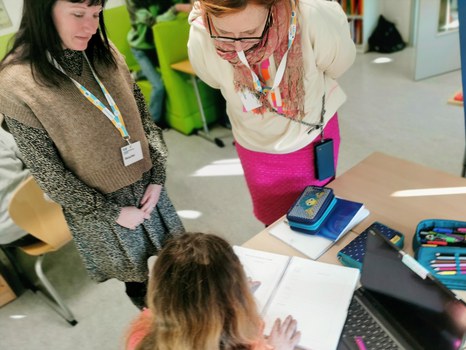 Blick von oben auf einen Schreibtisch. Eine Schülerin zeigt zwei ALSO-Teilnehmerinnen ihr Logbuch.