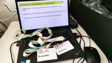 Ein Laptop zeigt die Folie "Austausch zum aktuellen Projektstand". Auf der Tastatur liegen Besucher-Schilder der Universitätsschule Dresden.