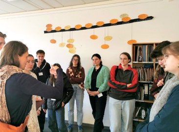 10 Studierende bei der Schulführung in einem Lernraum. Sie stehen vor einem Materialregal und unter einem Zeitstrahl. Sie schauen zur Projektmitarbeitern, die erklärt.