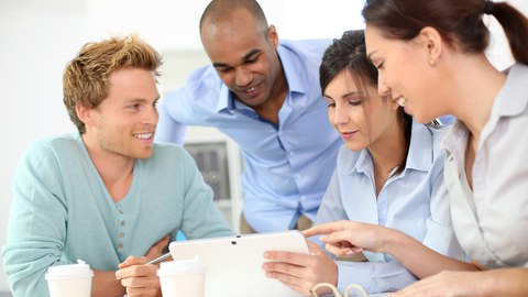 Das Foto zeigt vier Studierende, die gemeinsam auf ein Tablet schauen. Die Studentin am rechten Bildrand zeigt auf etwas und alle Studierenden lächeln.