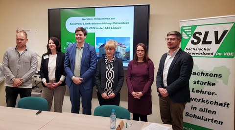 6 Personen stehen in formeller Kleidung vor einem großen Bildschirm und einem Banner bei der Konferenz Lehrkräfteausbildung in Ostsachsen des SLV