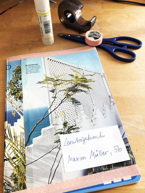 Mit buntem Papier beklebtes Lerntagebuch von Schüler Maxi Müller, Klasse 5 c. Im Hintergrund liegen Schere, Leim und Klebeband
