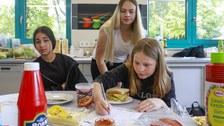 Eine Studentin und zwei Schülerinnen arbeiten in der Schulküche der Richard-von-Schlieben-Oberschule in Zittau an einem Projekt zum Thema Ernährung. Auf dem Tisch stehen Zutaten für ein Sandwich und einfertiges Essen. Eine Schülerin bearbeitet ein Aufgabe