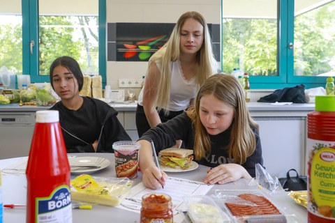 Eine Studentin und zwei Schülerinnen arbeiten in der Schulküche der Richard-von-Schlieben-Oberschule in Zittau an einem Projekt zum Thema Ernährung. Auf dem Tisch stehen Zutaten für ein Sandwich und einfertiges Essen. Eine Schülerin bearbeitet ein Aufgabe