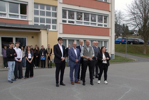 Vor einer Plattenbauschule steht eine Gruppe Erwchachsener, dahinter vereinzelte Schülerinnen und Schüler.