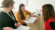 Projektleiterin Anke Langner im Gespräch mit Schulleiterin Sabrina Hälschke aus Zittau und einer weiteren Frau. Sie blicken in Logbücher der Universitätsschule Dresden.