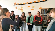 10 Studierende bei der Schulführung in einem Lernraum. Sie stehen vor einem Materialregal und unter einem Zeitstrahl. Sie schauen zur Projektmitarbeitern, die erklärt.