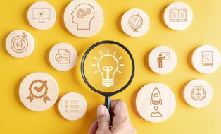 Foto einer Glühbirne, die unter einer Lupe betrachtet wird. Um die Glühbirne herum liegen rund Holzplättchen mit verschiedenen Icons wie ein Globus, eine Rakete oder ein Buch. 