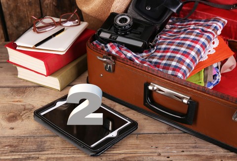 Das Foto zeigt einen braunen Koffer. In dem Koffer befinden sich Kleidung und eine alte Kamera. Davor liegt ein Tablet auf dem die Zahl 2 steht. Außerdem erkennt man Bücher, ein Notizbuch und eine Brille.