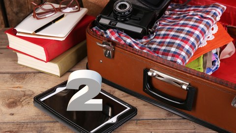 Das Foto zeigt einen braunen Koffer. In dem Koffer befinden sich Kleidung und eine alte Kamera. Davor liegt ein Tablet auf dem die Zahl 2 steht. Außerdem erkennt man Bücher, ein Notizbuch und eine Brille.