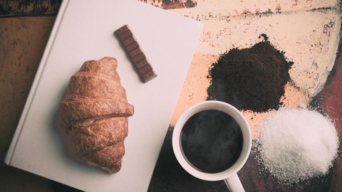 Buch mit einem Croissant und einem Schokoriegel drauf. Daneben steht ein Kaffee, Zucker und braunes Pulver.