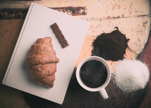 Buch mit einem Croissant und einem Schokoriegel drauf. Daneben steht ein Kaffee, Zucker und braunes Pulver.