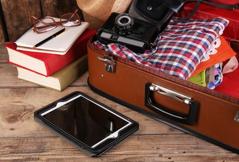 Das Foto zeigt einen braunen Koffer. In dem Koffer befinden sich Kleidung und eine alte Kamera. Davor liegt ein Tablet. Außerdem erkennt man Bücher, ein Notizbuch und eine Brille.