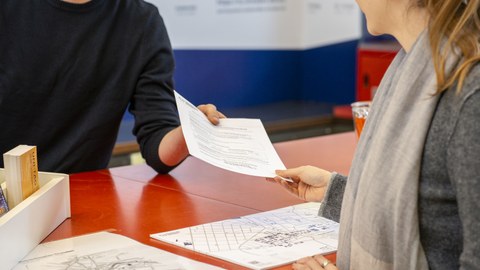 Foto von zwei Personen am SCS-Servicepoint der TU Dresden. Ein Mann reicht einer Frau ein Formular über den Tresen. 