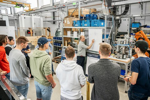 Foto eines Dozenten, der zwischen zwei Studierenden steht und ihnen ein Bauteil erklärt, das er in der Hand hält.